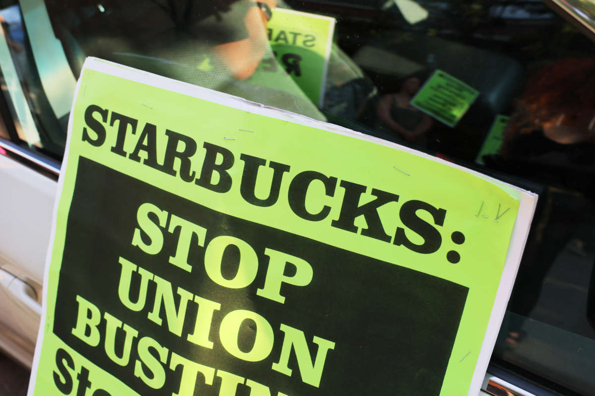 A sign reading: Starbucks: Stop Union Busting, leaned against a car with reflection of protesters in window