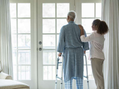 Caretaker assists elderly person, using a walker, as they stand at glass door looking at bright day