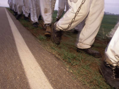 Shackled prisoners march in a chain gang as their overseer walks behind them