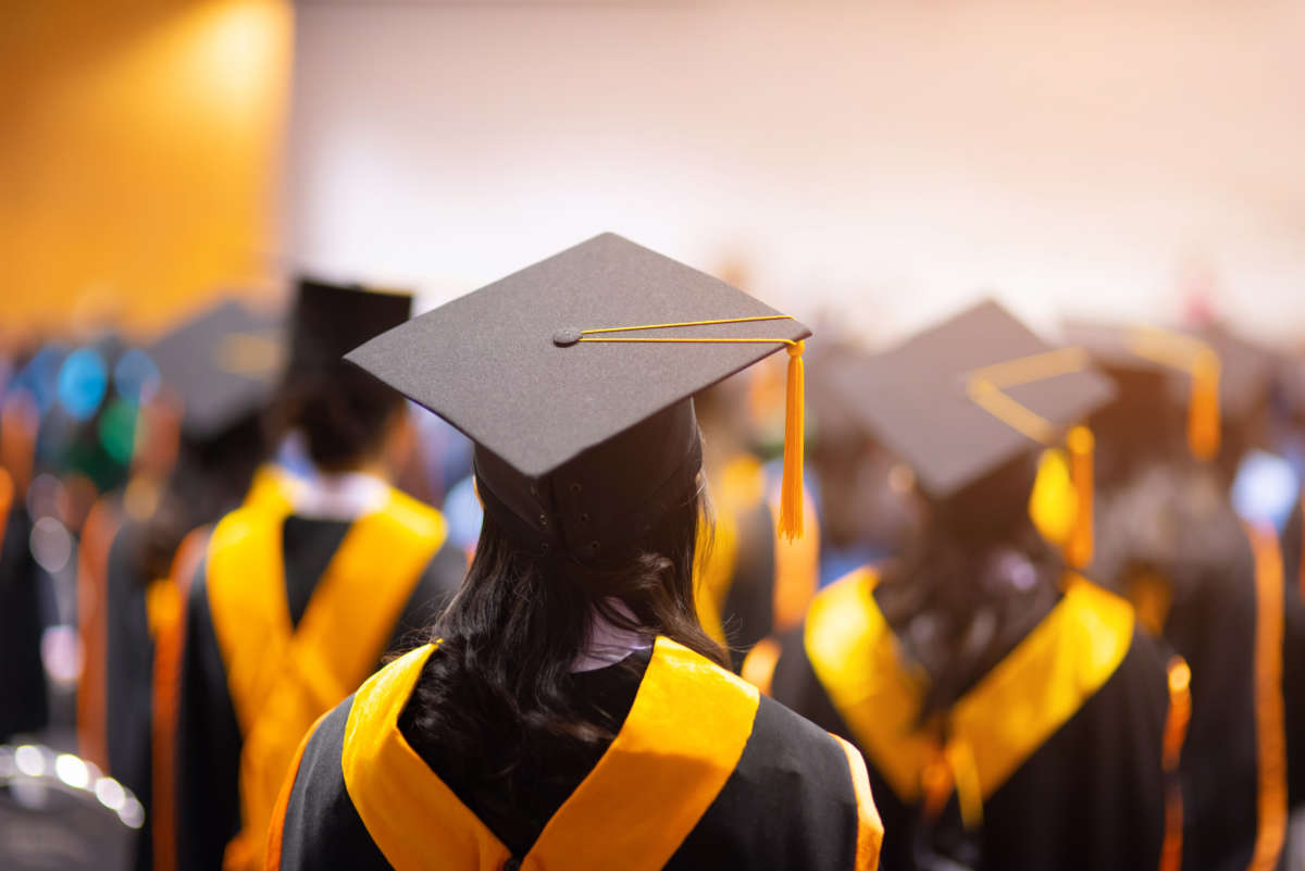 College graduates face away with, wearing black caps and gowns and yelllow sashes