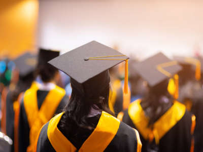 College graduates face away with, wearing black caps and gowns and yelllow sashes