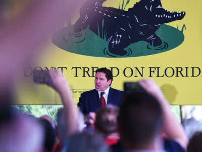 Florida Gov. Ron DeSantis speaks to supporters in front of large yellow 'don't tread on Florida' flag with crocodile illustration