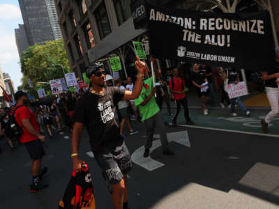 Christian Smalls, President of the ALU, leads pro-union protestors on a march down a New York street. Many carry signs, one prominent banner reads 'Amazon: Recognize the ALU!'