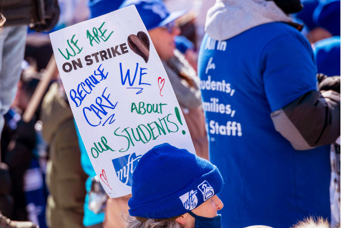 A person in blue cold-weather clothing participates in an outdoor protest while carrying a sign that reads "WE ARE ON STRIKE BECAUSE WE CARE ABOUT OUR STUDENTS"