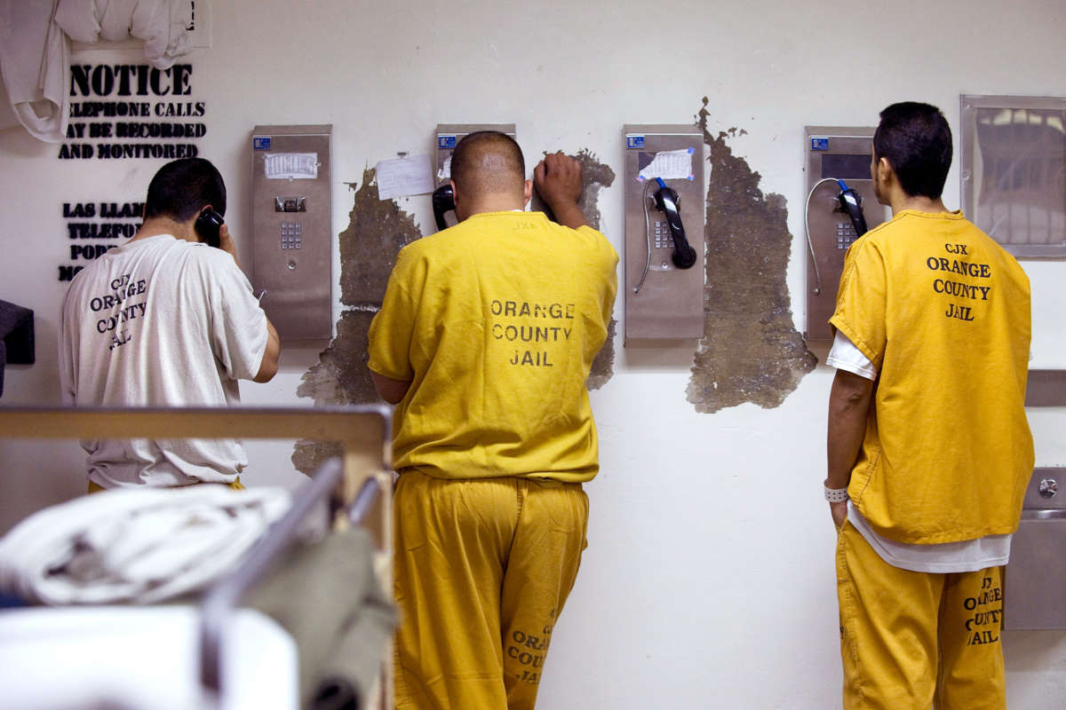 Prisoners in orange jumpsuits make phone calls against a damaged wall