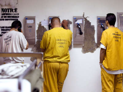 Prisoners in orange jumpsuits make phone calls against a damaged wall