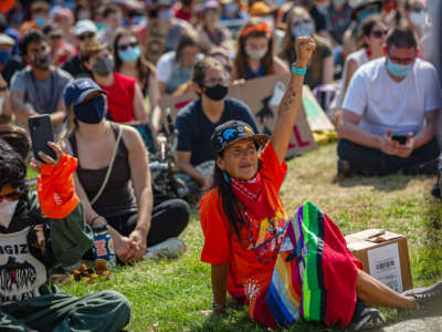 A woman raises a fist while sitting with other seated protesters at an outdoor event