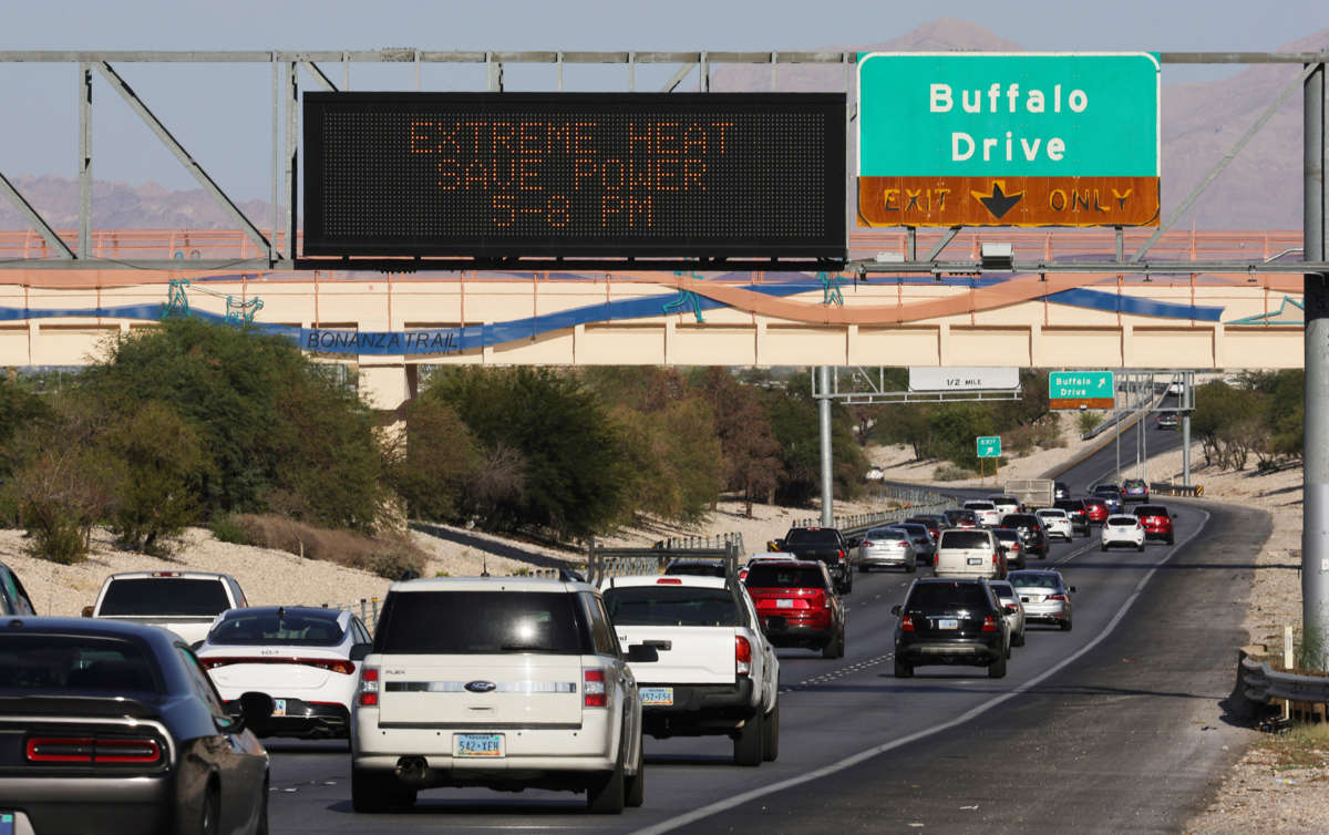 Cars drive on a highway with a lit sign reading "EXTREME HEAT - SAVE POWER 5-8 PM" hanging overhead