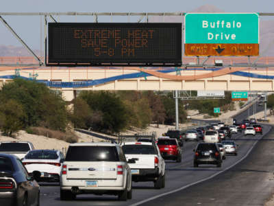 Cars drive on a highway with a lit sign reading "EXTREME HEAT - SAVE POWER 5-8 PM" hanging overhead