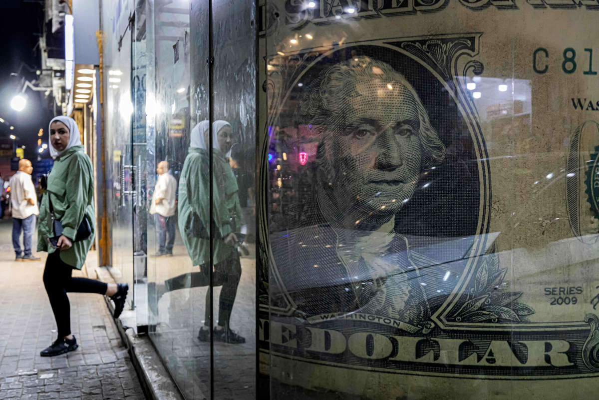 A hijabi exists a store with a large sign of a $1 bill prominently displayed in the window