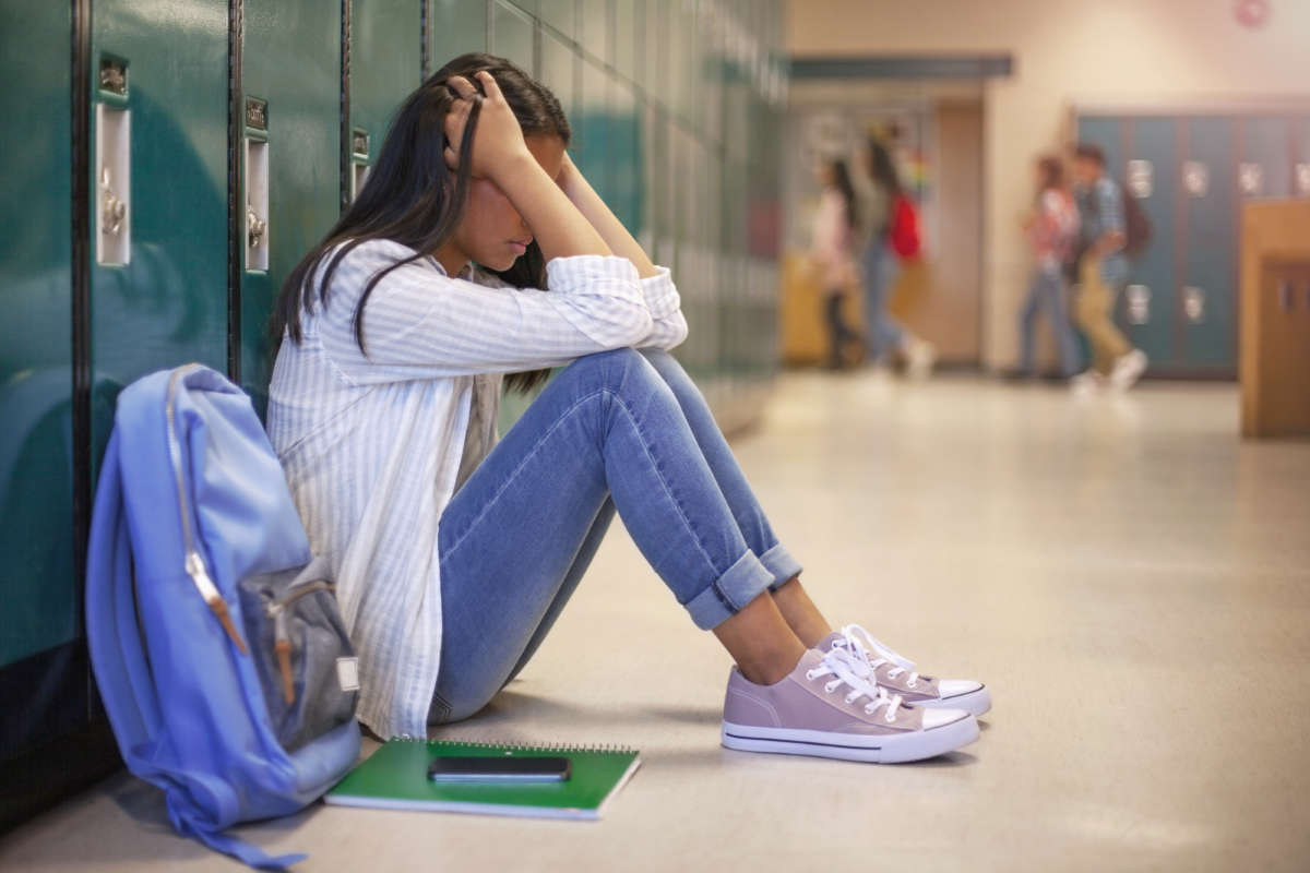 Frustrated teenage female student sitting with head in hands. She is sitting against metallic lockers.