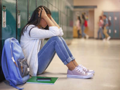 Frustrated teenage female student sitting with head in hands. She is sitting against metallic lockers.