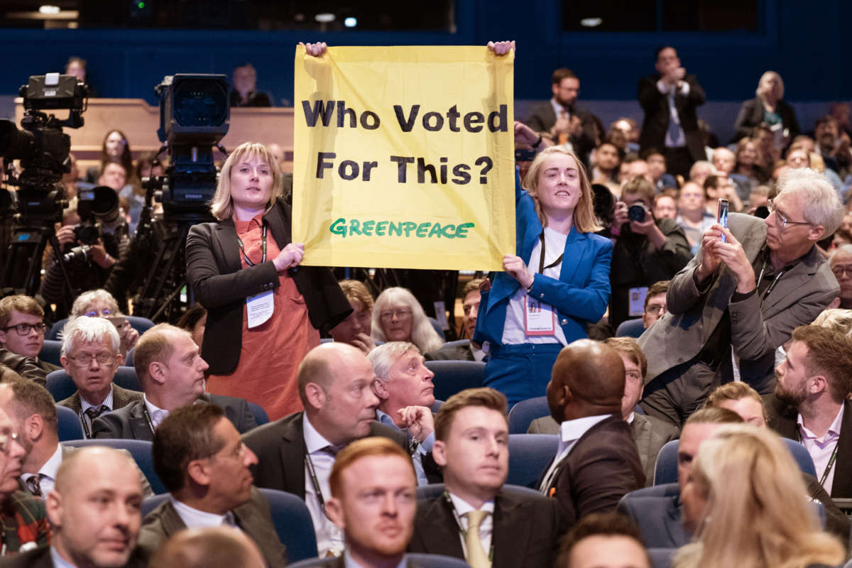 Two people hold up a banner reading "WHO VOTED FOR THIS" beneath which the Greenpeace wordmark is displayed