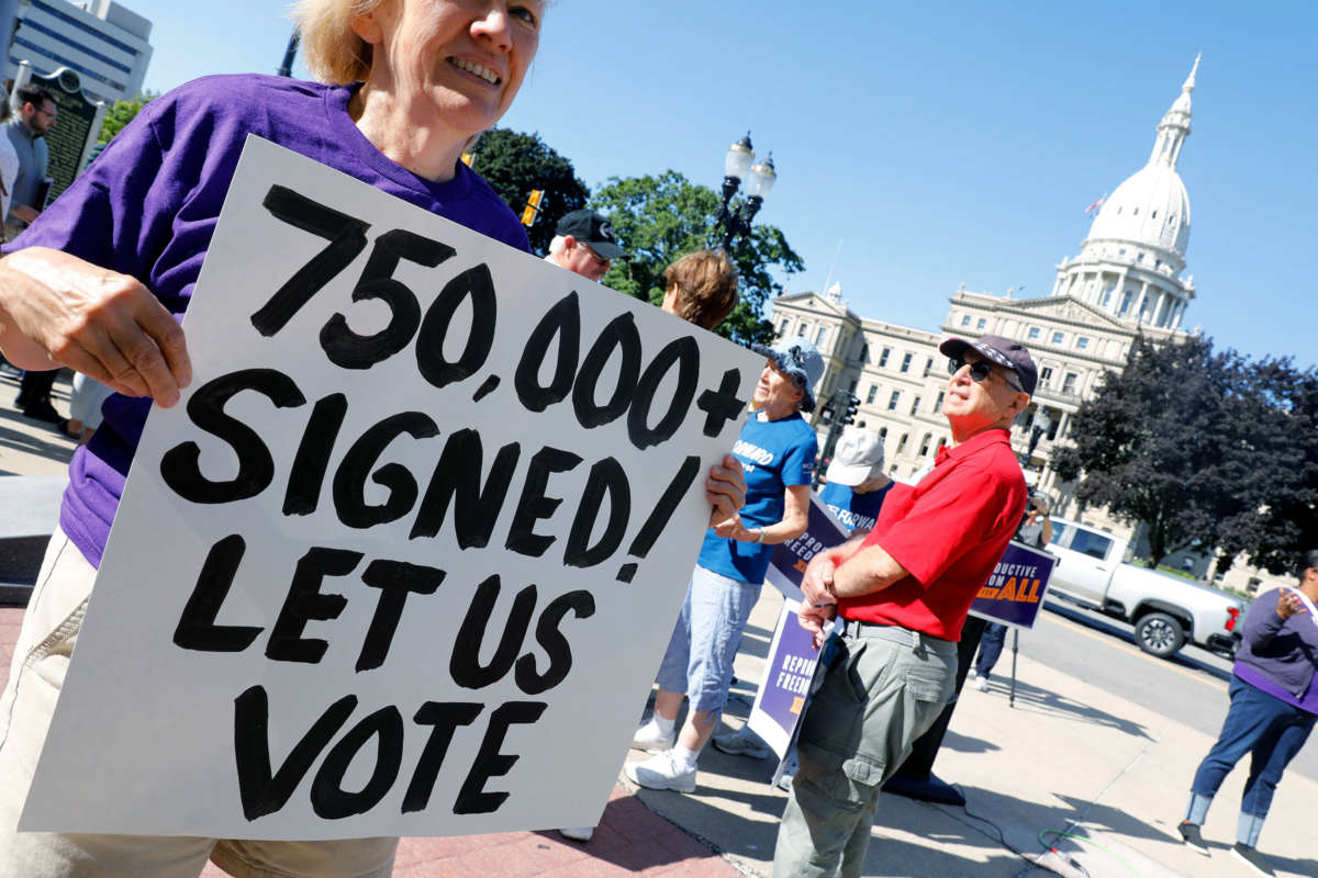 A protester in a purple shirt holds a sign reading "750,000+ SIGNED! LET US VOTE" outside of the Michigan State Capitol