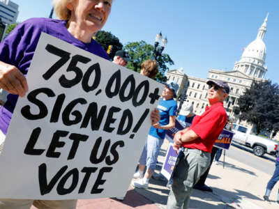 A protester in a purple shirt holds a sign reading "750,000+ SIGNED! LET US VOTE" outside of the Michigan State Capitol