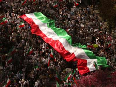 An aerial view of a massive protest wherein people march with a large banner of the Iranian flag