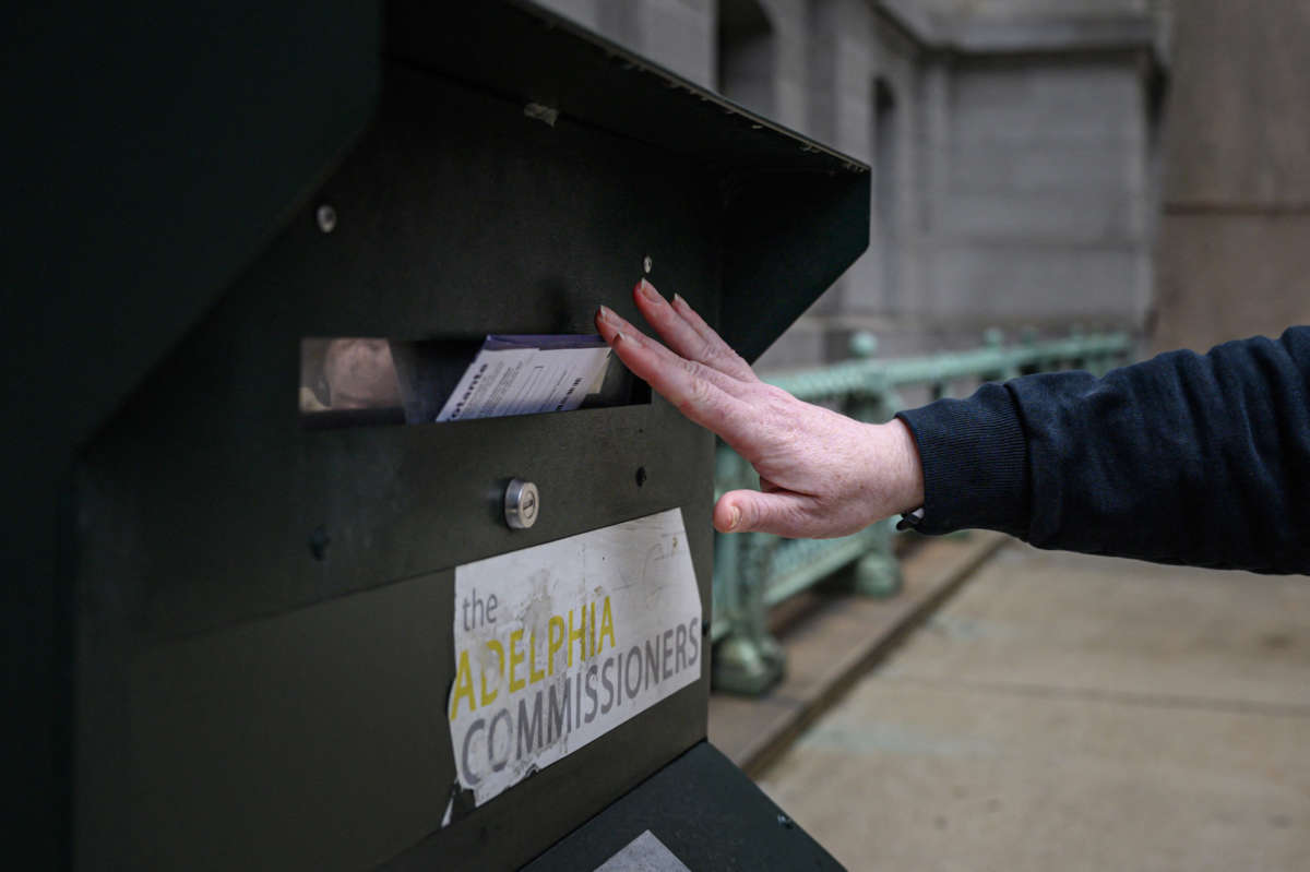 A person's hand is seen inserting a mail-in ballot into a dropbox