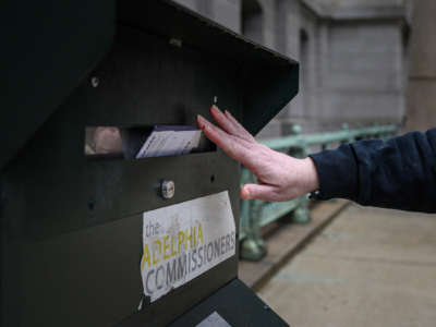 A person's hand is seen inserting a mail-in ballot into a dropbox