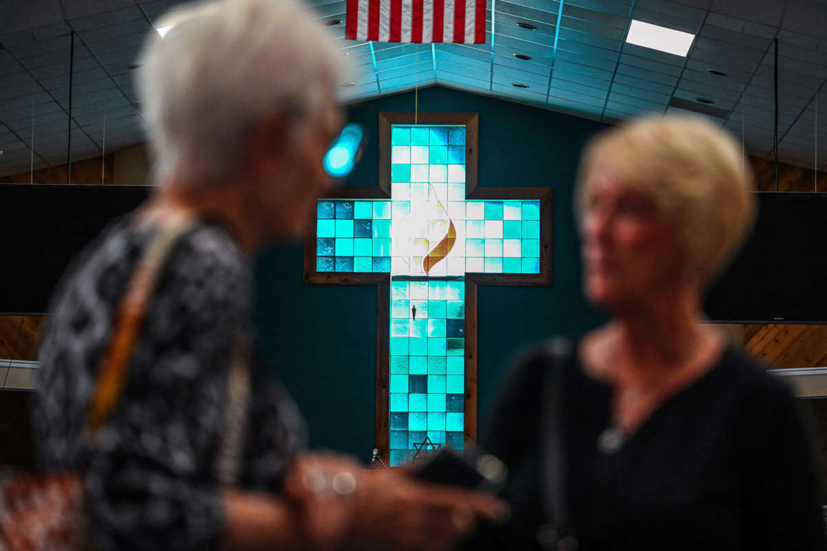 Two elderly women, out of focus, speak to eachother in front of a stained-glass window of a cross