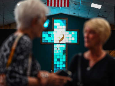 Two elderly women, out of focus, speak to eachother in front of a stained-glass window of a cross