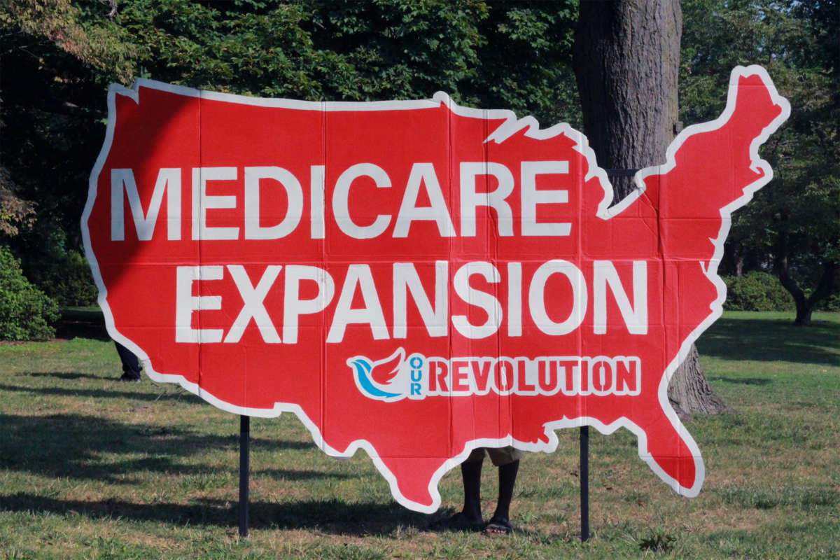 A cardboard sign in the shape of the contiguous continental United States reads "MEDICARE EXPANSION" with the "Our Revolution" logo beneath it