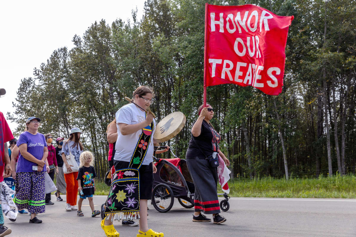 Water protectors, led by an elder carrying a flag reading "HONOR THE TREATIES" march down a wooded street