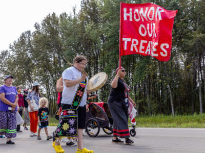 Water protectors, led by an elder carrying a flag reading "HONOR THE TREATIES" march down a wooded street