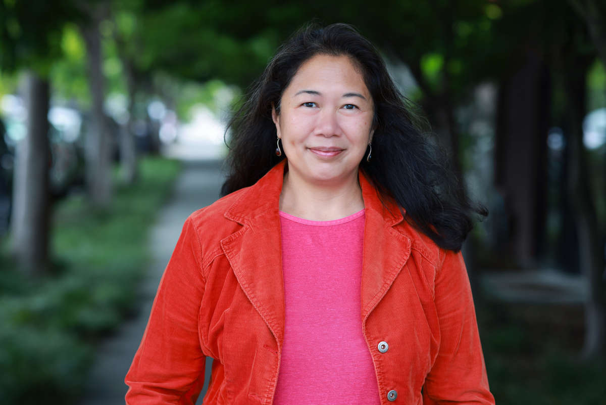 A woman stands on a wooded path, wearing a pink blouse with a red-orange jacket