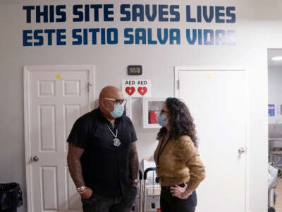 Two people in masks speak to oneanother in front of a wall with a painting reading "THIS SITE SAVES LIVES; ESTE SITIO SALVA VIDAS"