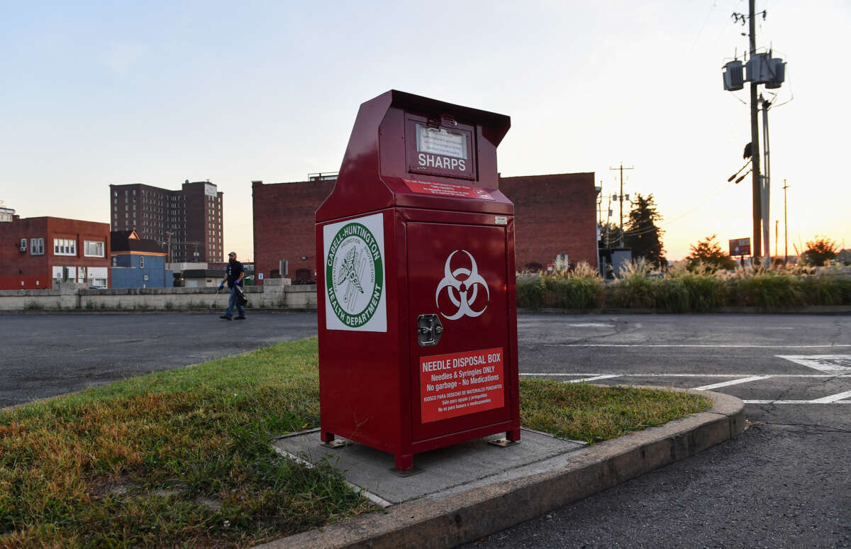 A red metal container labeled with the biohazard symbol and verbiage stating that it's to be used for sharps disposal sits in a vacant parking lot