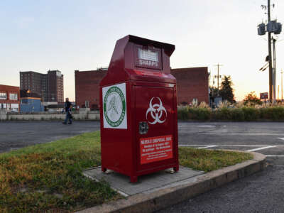 A red metal container labeled with the biohazard symbol and verbiage stating that it's to be used for sharps disposal sits in a vacant parking lot