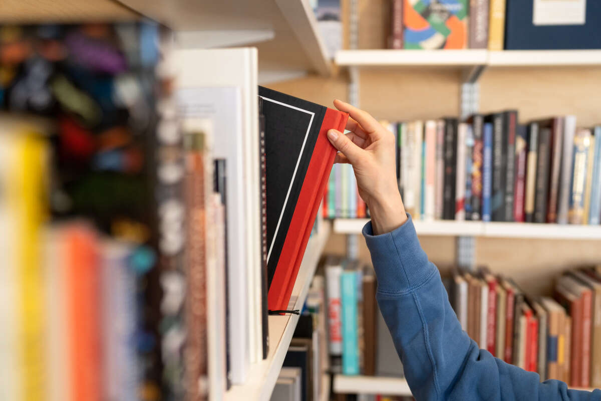 A woman's hand reaches for a black book with a red spine on a library shelf of assorted books.
