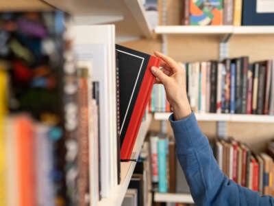 A woman's hand reaches for a black book with a red spine on a library shelf of assorted books.