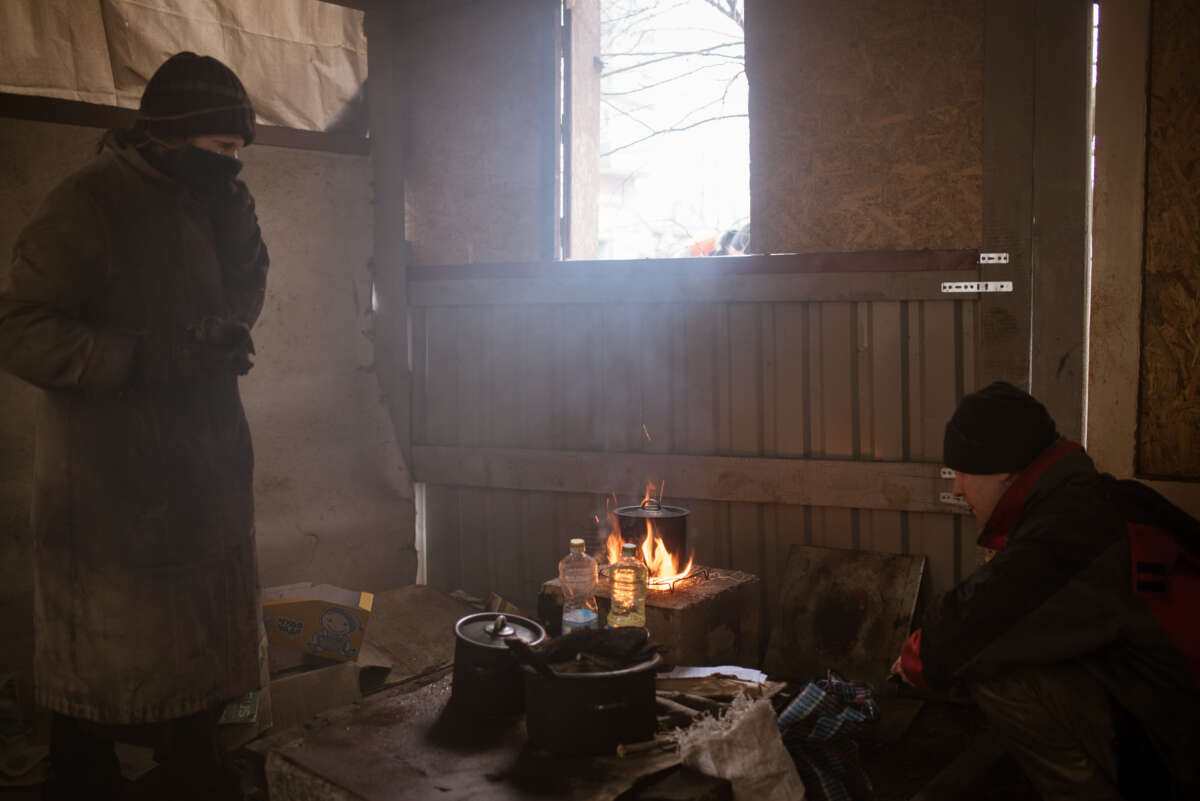 Two women dressed for the cold prepare lunch over a fire next to an open window