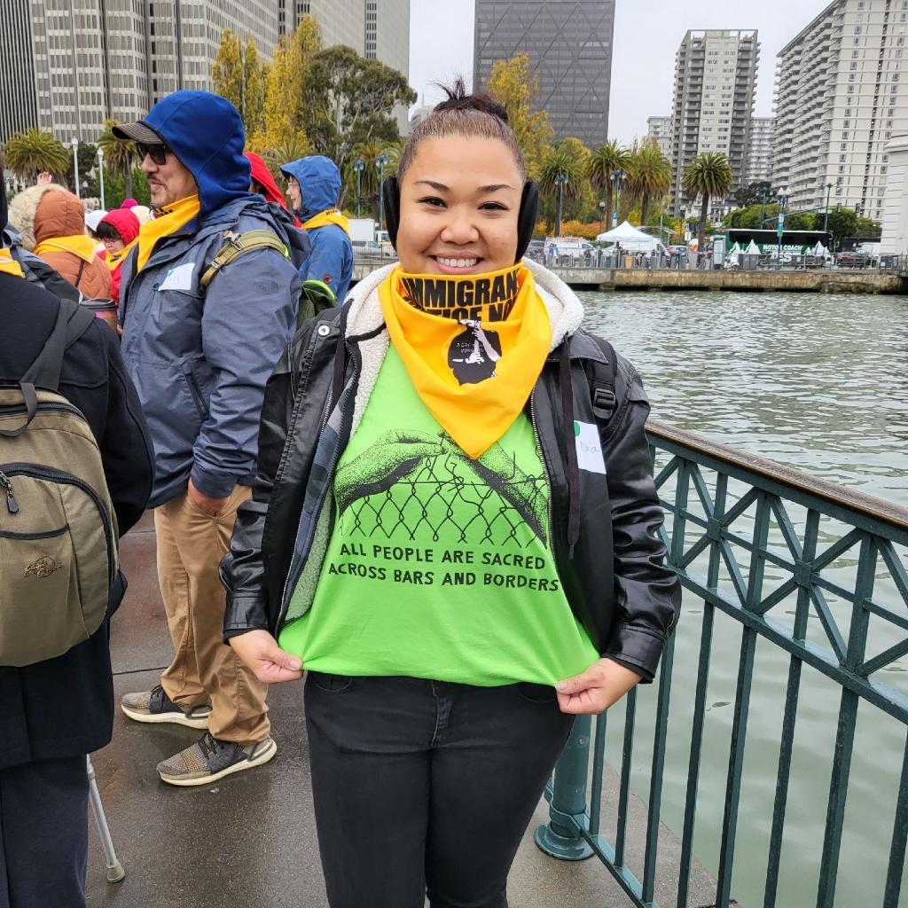 A woman in a group of other protesters displays the green shirt that she currently wears, reading: "ALL PEOPLE ARE SACRED ACROSS BARS AND BORDERS"