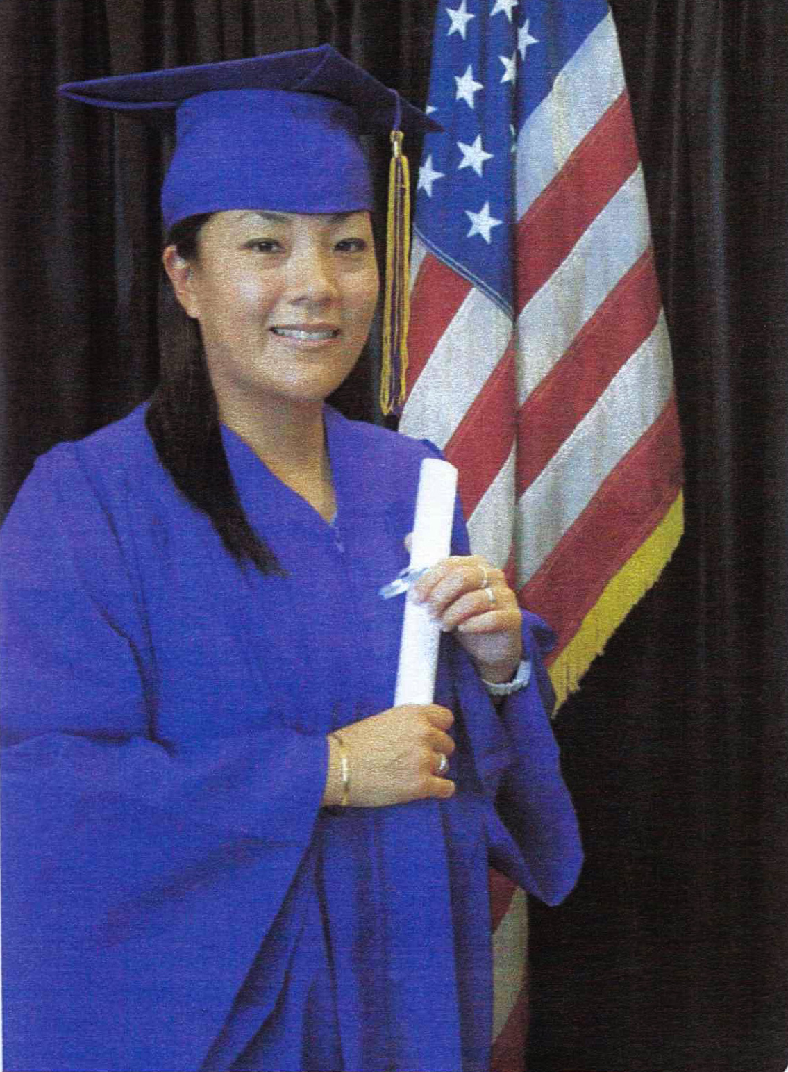 A woman in graduation robes and a mortarboard holds her diploma while standing in front of an American Flag.