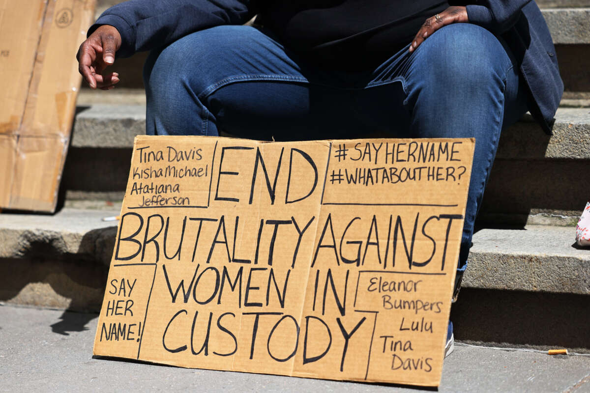 A seated protester rests behind a sign reading "END BRUTALITY AGAINST WOMEN IN CUSTODY" surrounded by the names of victims of the carcereal state