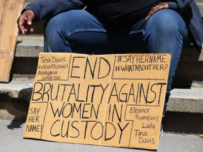 A seated protester rests behind a sign reading "END BRUTALITY AGAINST WOMEN IN CUSTODY" surrounded by the names of victims of the carcereal state