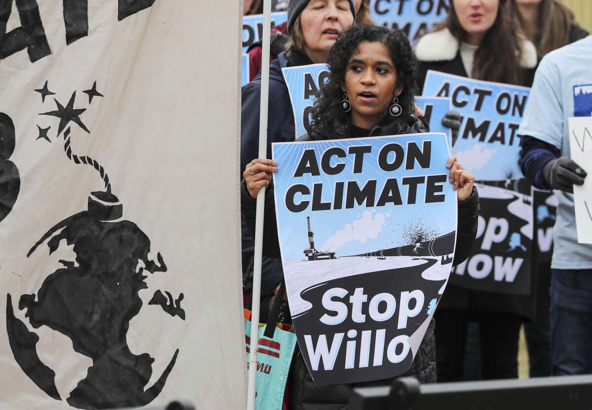 an activist holding a sign reading "ACT ON CLIMATE; STOP WILLOW" stands with others during an outdoor protest