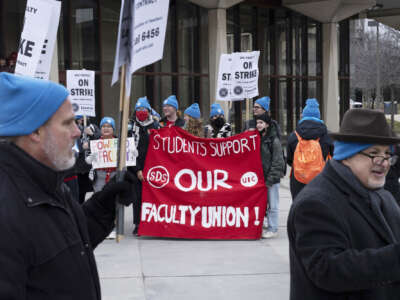 People hold a red banner reading "STUDENTS SUPPORT OUR FACULTY" as striking professors protest around them