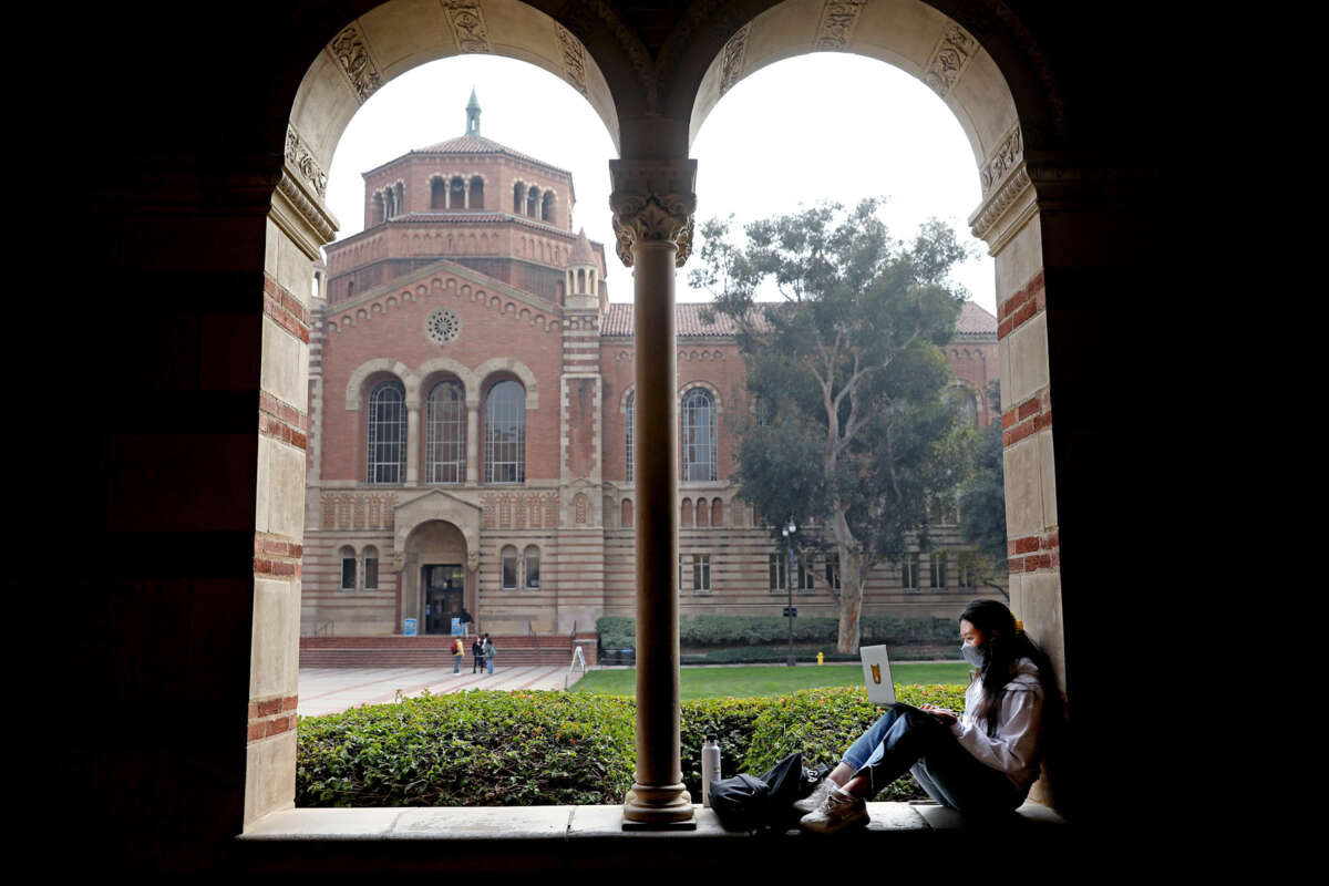 A masked student sits in one one of the arches of a university building to study on their lapotop