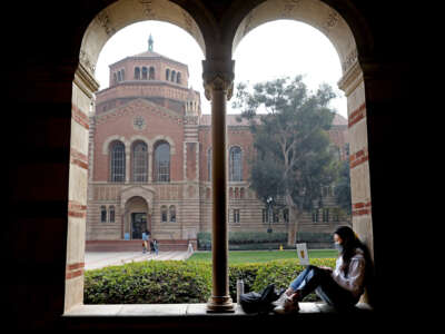 A masked student sits in one one of the arches of a university building to study on their lapotop