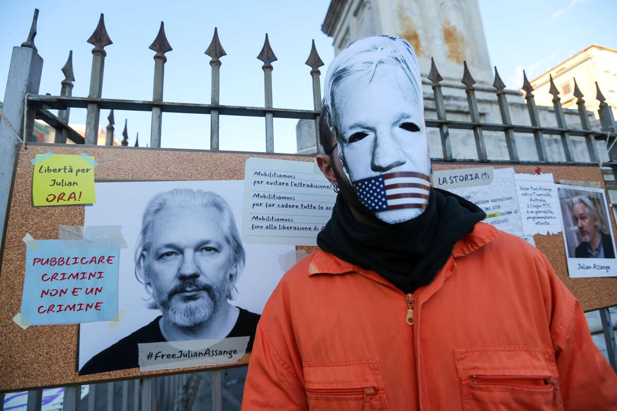 A protester in a mask made to resemble Julian Assange's face being gagged with the U.S. flag stands in front of a sign made in support of him