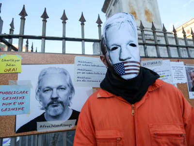 A protester in a mask made to resemble Julian Assange's face being gagged with the U.S. flag stands in front of a sign made in support of him