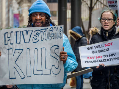 A man in blue holds a sign reading "HIV STIGMA KILLS" while another behind him holds a sign reading "WE WILL NOT GO BACK; SAY NO TO HIV CRIMINALIZATION" during an outdoor protest