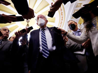 A masked Bernie Sanders speaks into recording devicies held up by reporters surrounding him