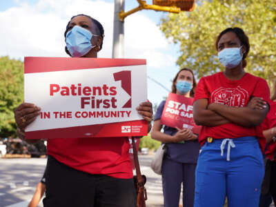 Masked nurses, one of whom holds a sign reading "PATIENTS FIRST IN THE COMMUNITY," participate in an outdoor protest