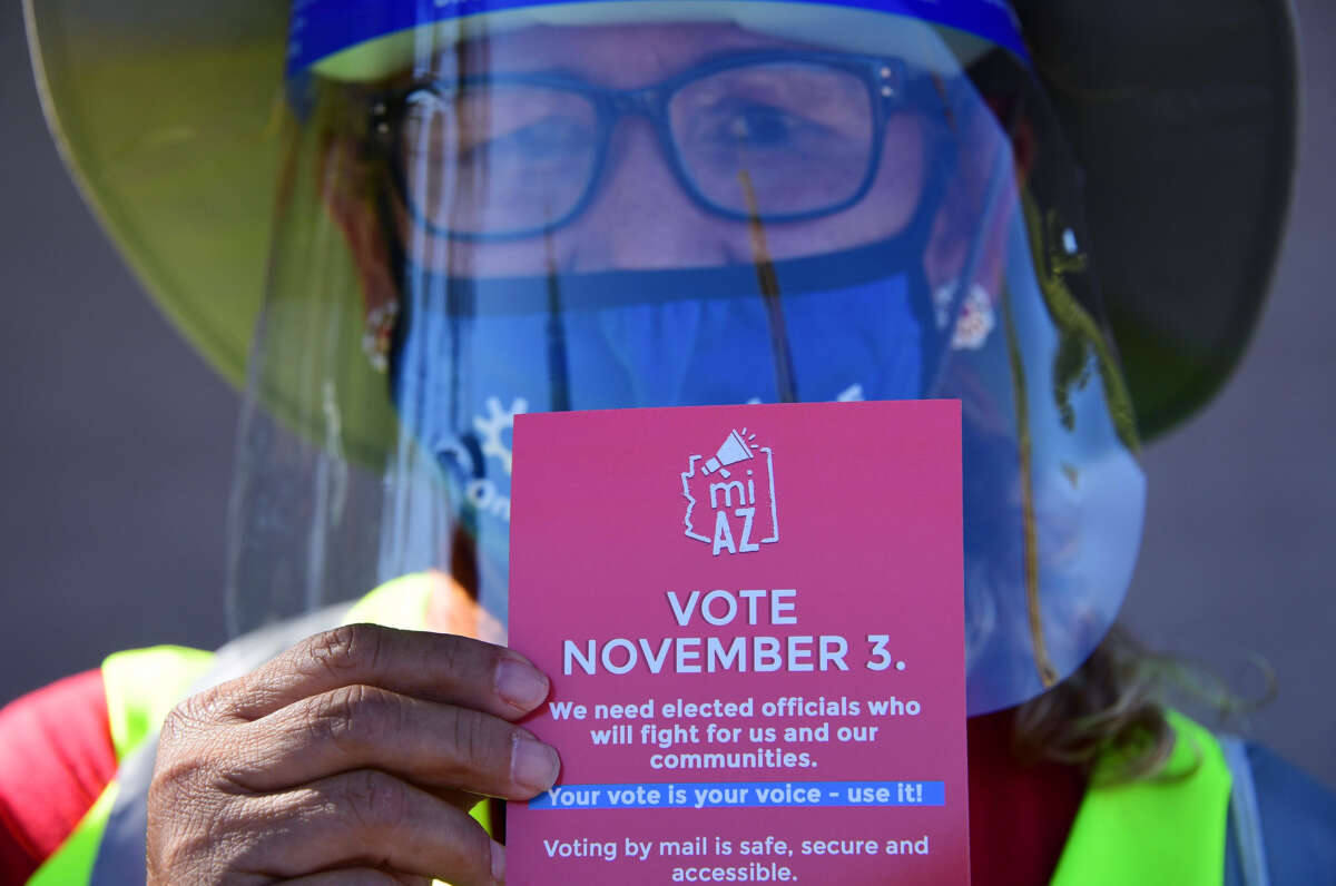 An election woker wearing a face sheild and visibility gear holds out a pamphlet urging people to vote