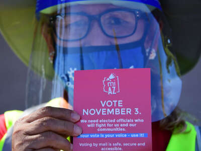 An election woker wearing a face sheild and visibility gear holds out a pamphlet urging people to vote