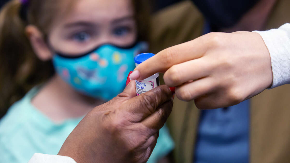 A girl in a mask watches an adult hand over a vaccine vial to another adult